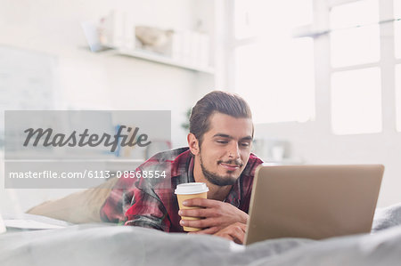 Young man drinking coffee at laptop on bed