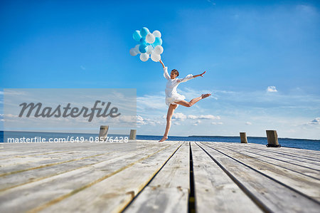 Young woman dancing on wooden pier, holding bunch of balloons