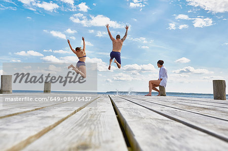 Young woman sitting on post on wooden pier, watching friends as they jump into sea