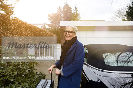 Woman near car on driveway holding suitcase looking at camera smiling