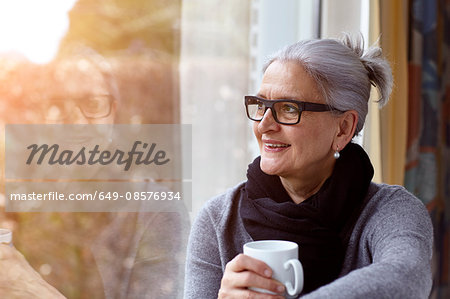 Woman wearing glasses holding coffee cup looking out of window smiling