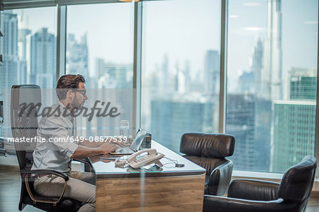 Businessman using laptop at desk with window view of Burj Khalifa, Dubai, United Arab Emirates