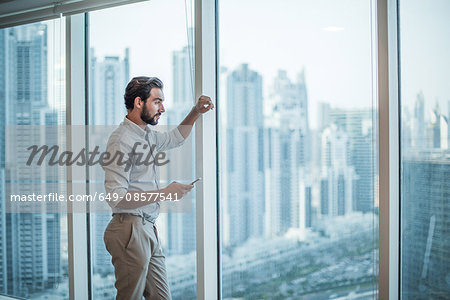 Businessman with smartphone staring through window with skyscraper  view, Dubai, United Arab Emirates