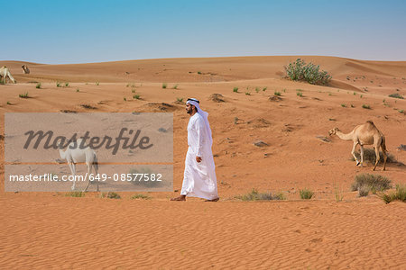 Middle eastern man wearing traditional clothes walking past camels in desert, Dubai, United Arab Emirates