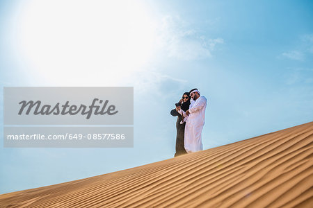 Middle eastern couple wearing traditional clothes taking smartphone selfie on desert dune, Dubai, United Arab Emirates