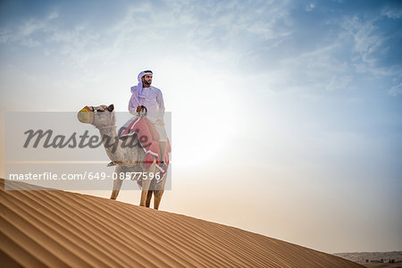 Man wearing traditional middle eastern clothes riding camel in desert, Dubai, United Arab Emirates