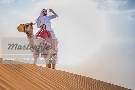 Man wearing traditional middle eastern clothes riding camel in desert, Dubai, United Arab Emirates