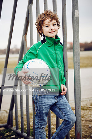 Boy leaning against railings holding football looking at camera smiling