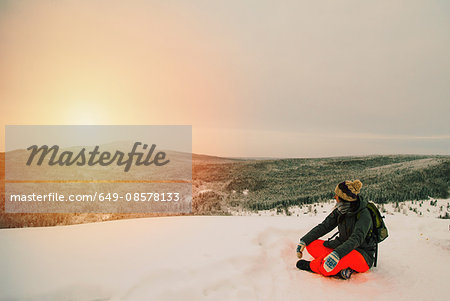 Woman on snow covered landscape sitting looking at view of sunset over mountain