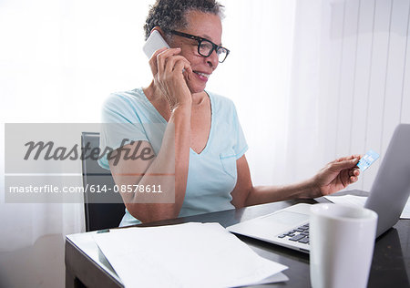 Senior woman sitting at table, using laptop, paperwork on table
