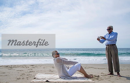 Senior couple on beach, man taking photograph of woman using smartphone