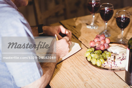 Close up view of man  writing in a book while tasting red wine and eating cheese