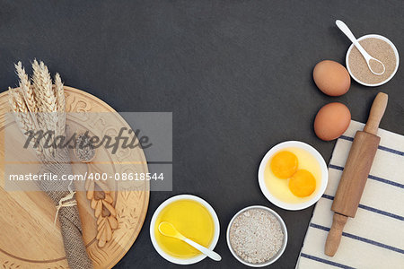Bread baking ingredients and utensils with wheat sheaths and grain in a love spoon on grey slate background.