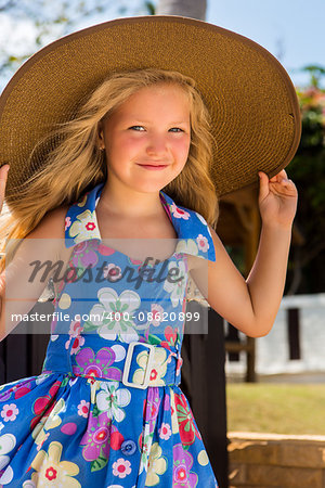 Outdoor portrait of beautiful girl in blue floral dress and beach straw hat. She holds her big hat and smile to camera. Summer sunny day. House with wooden fence at tropical background. Mothers day.