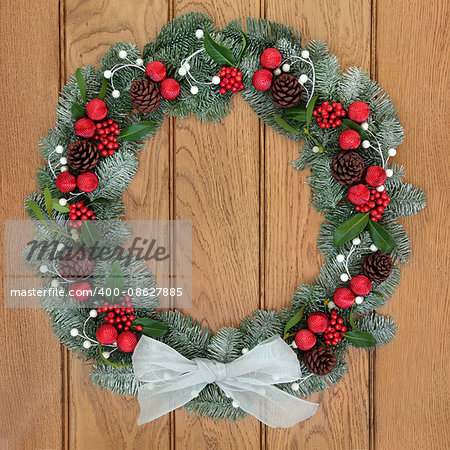 Christmas wreath with white bow, red bauble decorations, holly, pine cones, mistletoe and snow covered blue spruce fir over oak wood front door background.