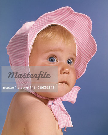 1960s BABY GIRL WITH UNCERTAIN FACIAL EXPRESSION WEARING PINK AND WHITE CHECKED BONNET TIED IN BOW UNDER CHIN