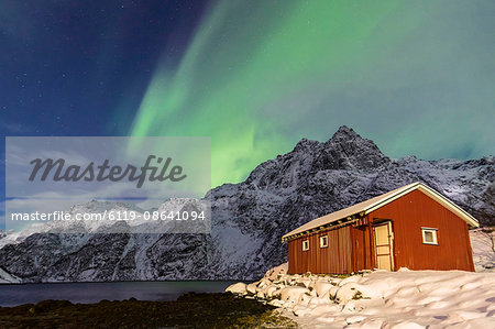 Northern Lights (aurora borealis) illuminate snowy peaks and the wooden cabin on a starry night at Budalen, Svolvaer, Lofoten Islands, Arctic, Norway, Scandinavia, Europe