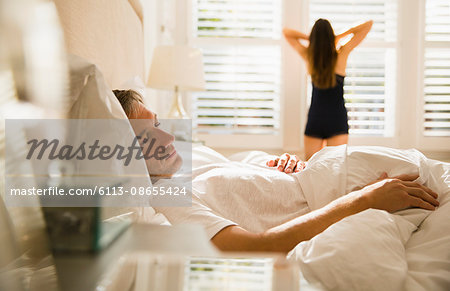 Wife stretching at morning window behind husband sleeping in bed