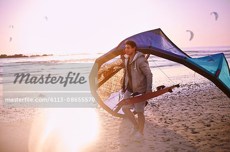 Man carrying kiteboarding equipment on sunset beach