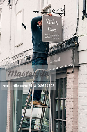 A man on a ladder fixing a painted name sign onto a bracket on a shopfront.