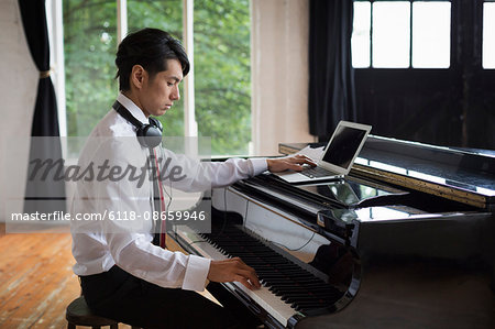 Young man sitting at a grand piano in a rehearsal studio, playing and simultaneously using a laptop computer.