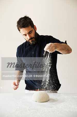 Baker standing at a table, liberally sprinkling flour over bread dough.