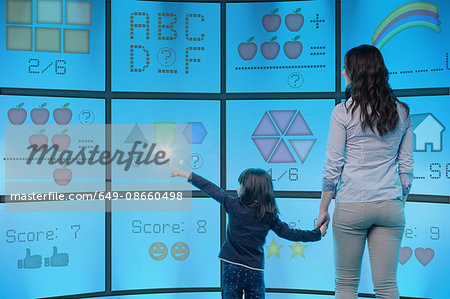 Mother and daughter standing in front of graphical screens showing educational images, girl reaching out to touch screen
