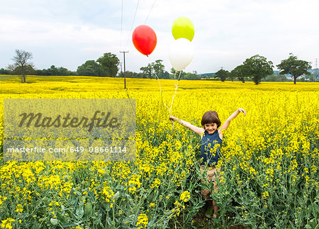 Boy standing in yellow flower field holding red, yellow and white balloons