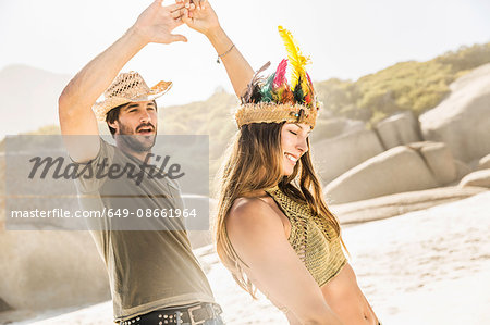 Mid adult couple wearing straw hat and feather headdress dancing on beach, Cape Town, South Africa