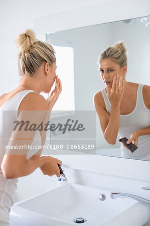 Beautiful young woman applying moisturizer on her face in bathroom