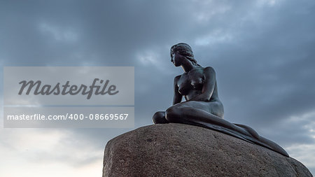 Low angle view of Little Mermaid statue on large boulder looking away in Denmark under cloudy sky