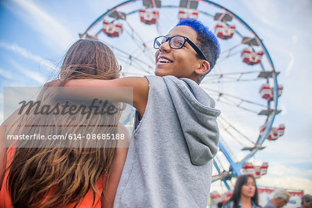 Rear view of teenage couple looking over shoulder in front of ferris wheel in amusement park