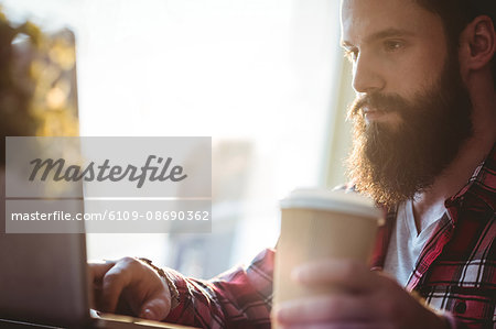 Hipster with disposable cup and laptop sitting by window at cafe