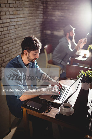 Handsome young man working on laptop with customer in background at cafe