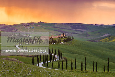 Valdorcia, Siena, Tuscany, Italy. Road of cypresses leading to a farmhouse with a stormy sunset in the background.
