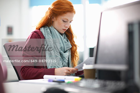 Young female college student at computer desk reading paperwork