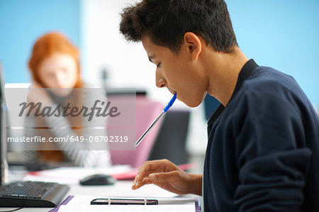 Young male college student at computer desk calculating on smartphone