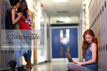 Young female college student sitting on locker room floor reading whilst typing on laptop