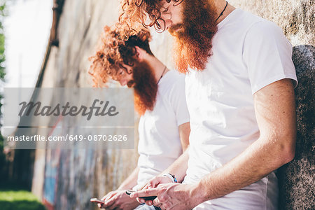 Young male hipster twins with red beards leaning against wall texting on smartphones