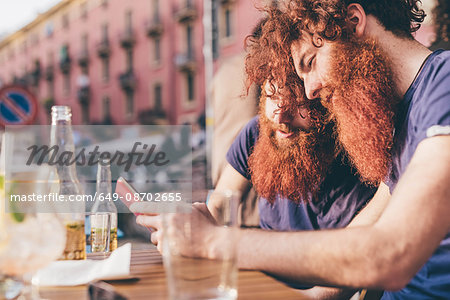 Young male hipster twins with red hair and beards reading smartphone texts at sidewalk bar
