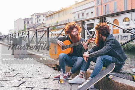 Young male hipster twins with red hair and beards playing guitar on canal waterfront