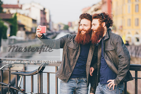 Young male hipster twins with red hair and beards taking smartphone selfie on canal waterfront