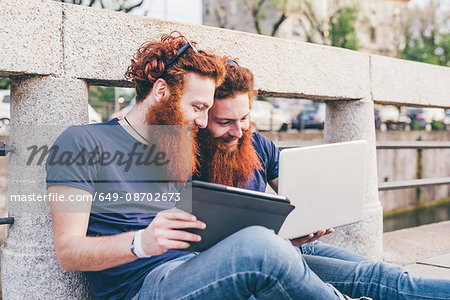 Young male hipster twins with red hair and beards sitting on bridge browsing digital tablet