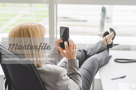 Mature businesswoman with feet up on office desk using smartphone touchscreen
