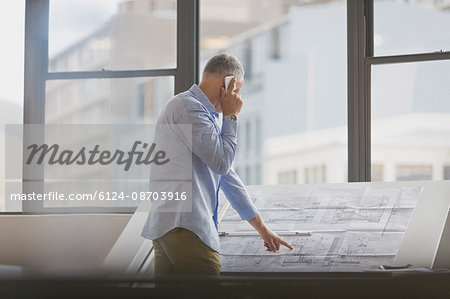 Architect talking on cell phone reviewing blueprints in office