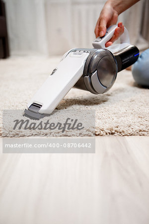 Female hand holding small white cordless vacuum cleaner and cleaning rug indoors