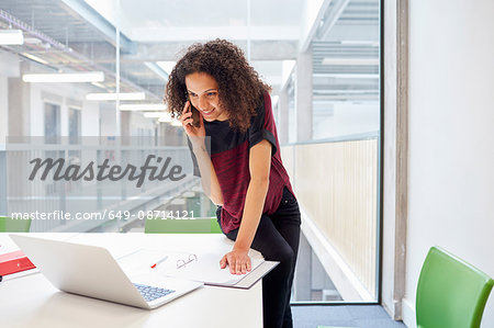 Female designer reading laptop and talking on smartphone  in design studio