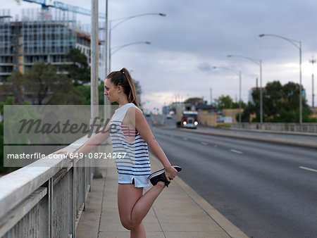 Young female runner stretching legs on highway bridge at dawn