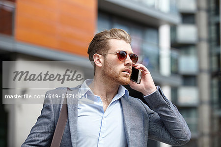 Young businessman wearing sunglasses talking on smartphone outside office, London, UK