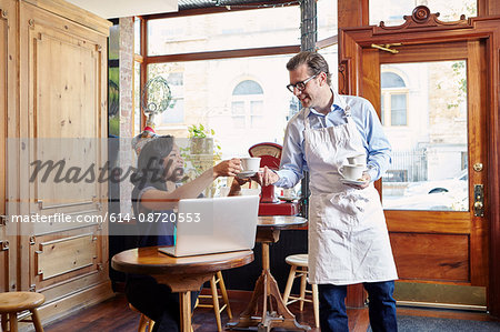 Young woman sitting in cafe, using laptop, male worker taking empty coffee cup